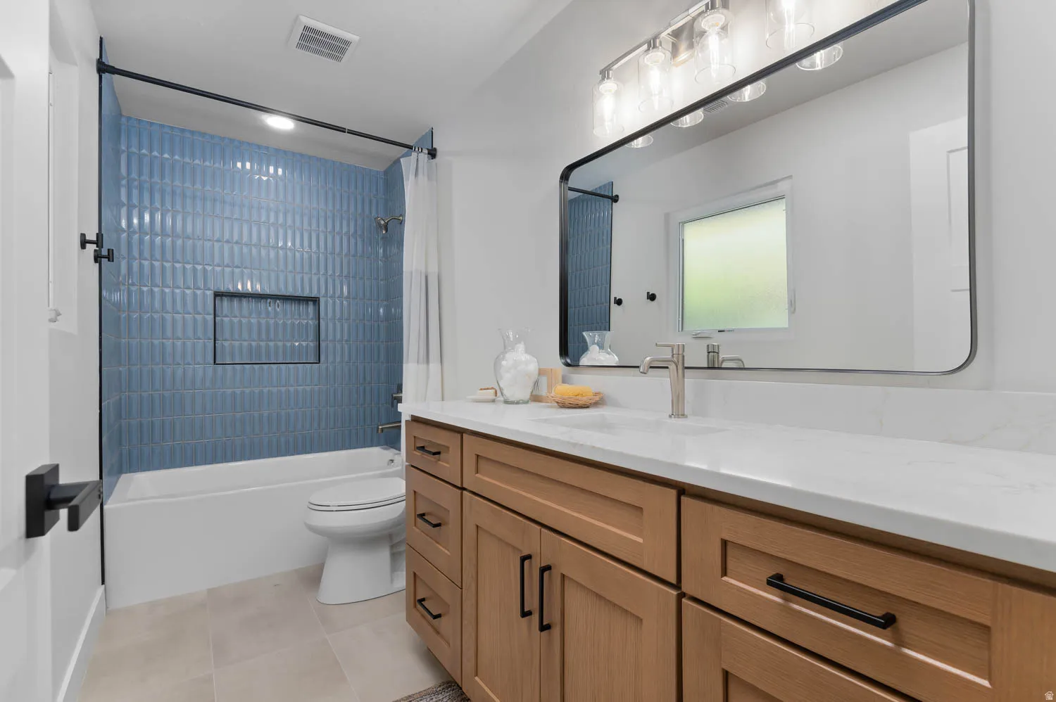 Bathroom featuring a wood-finish double vanity with quartz countertop, a rectangular framed mirror with brushed nickel vanity lighting, a tub/shower combination with blue textured tile surround and recessed niche, and light-colored floor tiling