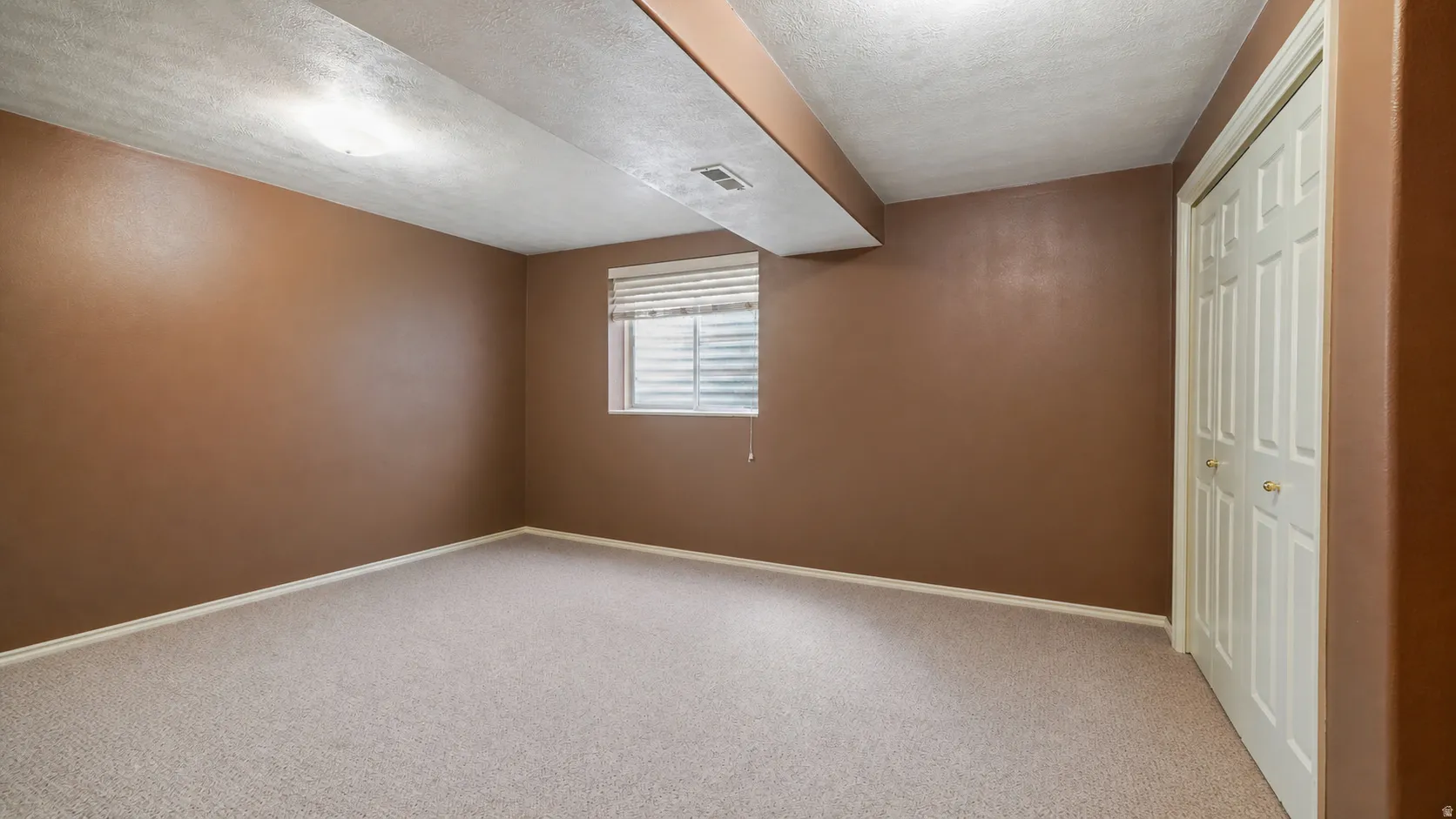 Carpeted room featuring a window with horizontal blinds, a white bi-fold closet door, and a painted ceiling beam