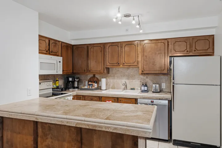 Kitchen with white appliances, a peninsula, wood finish cabinets, backsplash, and tile counters