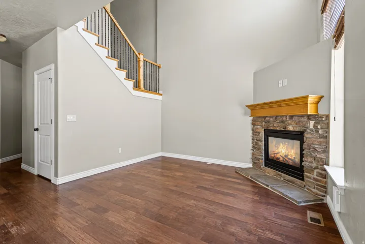 Spacious living area featuring wood-finish flooring, a stone-faced fireplace with a substantial wooden mantel, and a multi-level staircase with decorative metal balusters