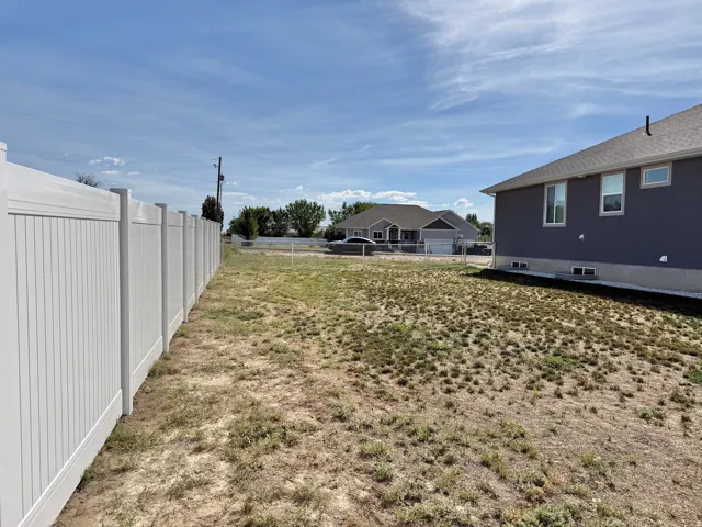 White privacy fence, exterior siding, multiple windows, gravel perimeter, and undeveloped ground cover