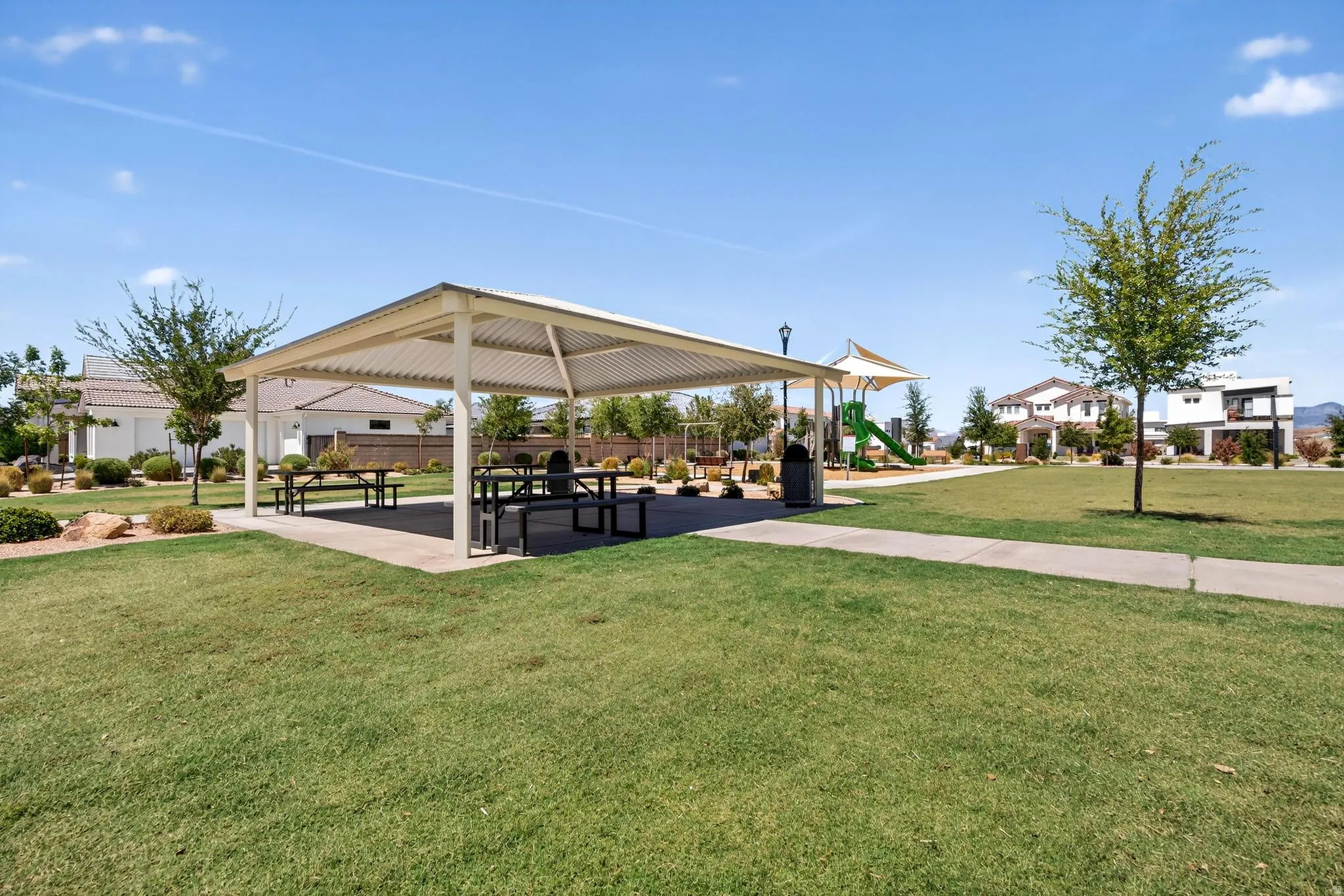 Community park featuring a large covered picnic area with multiple tables, a playground with slides and climbing elements, mature landscaping, and an expansive lawn