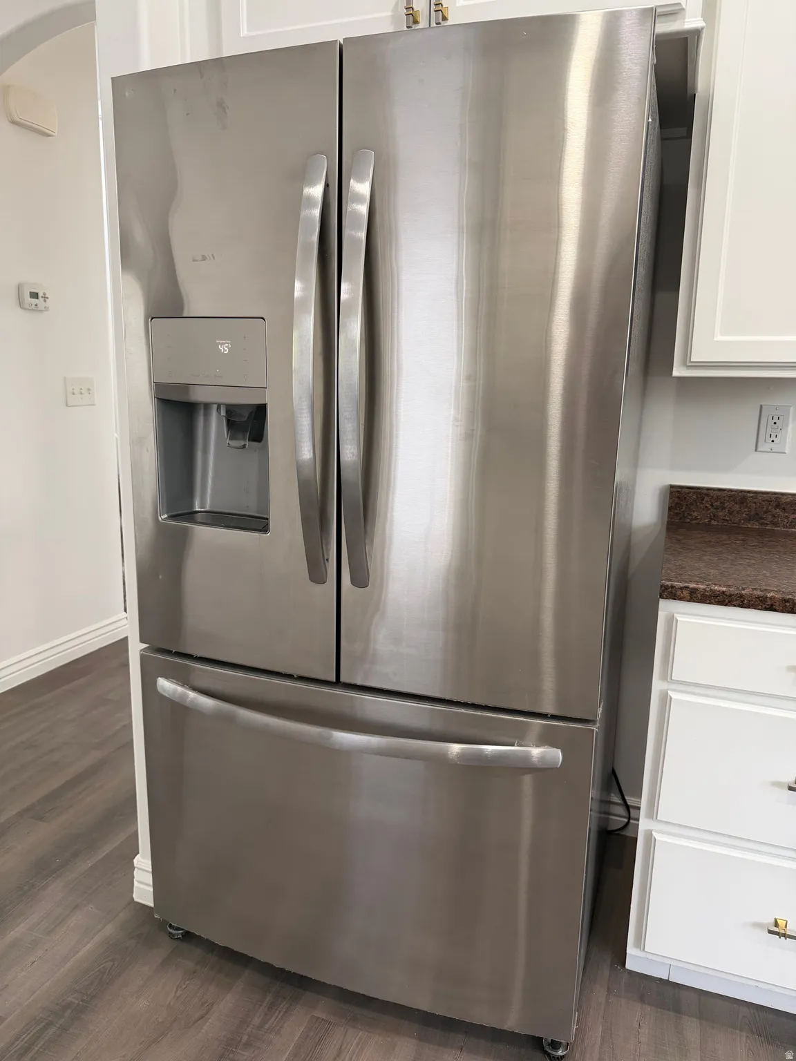 Kitchen view of stainless steel fridge with ice dispenser, dark countertops, white cabinets, and dark wood-style floors