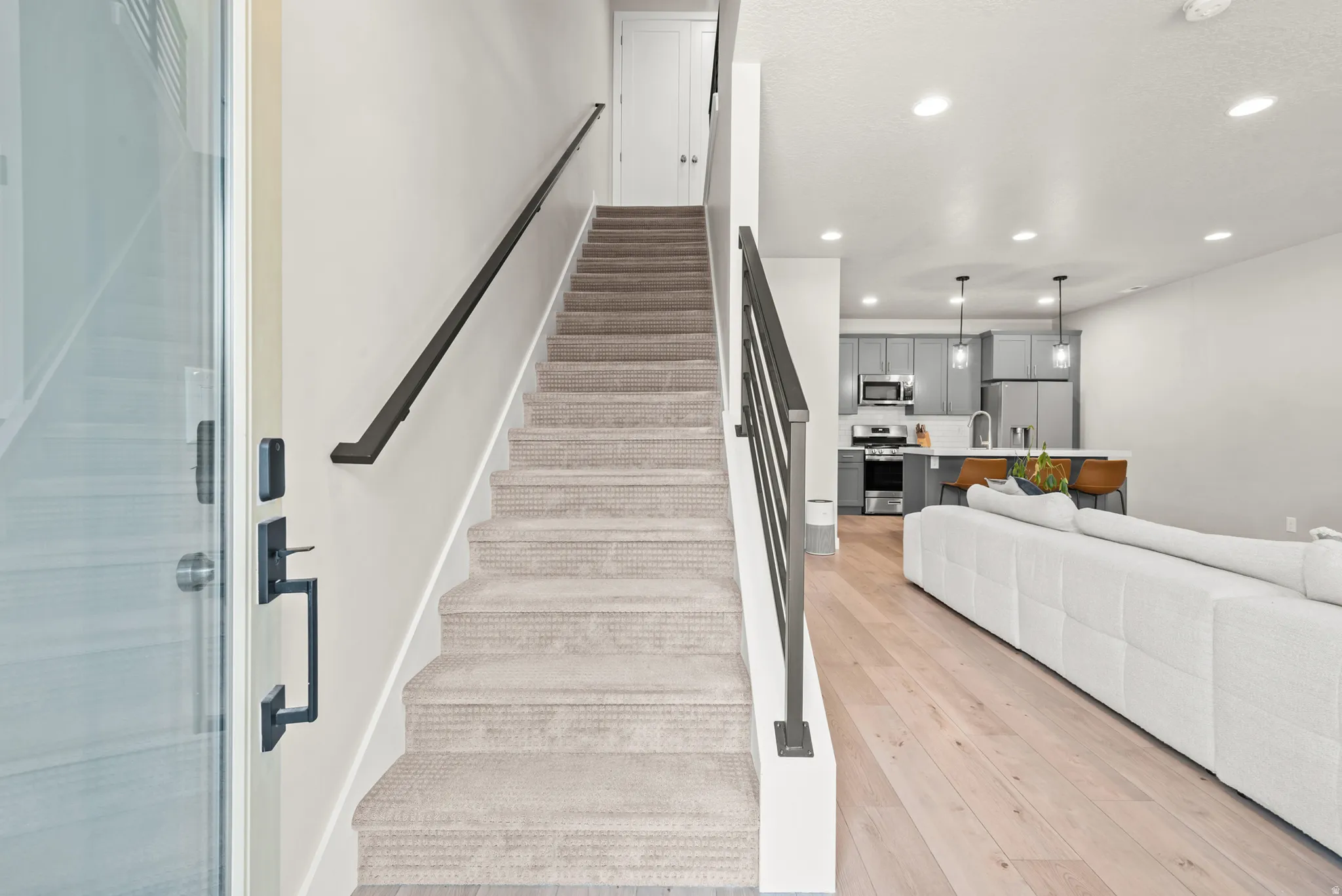 Open-concept living area featuring light wood-finish flooring, recessed lighting, a modern staircase with metal balusters and handrail, a kitchen with gray cabinetry, and stainless steel appliances