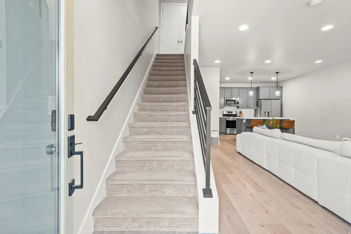 Open-concept living area featuring light wood-finish flooring, recessed lighting, a modern staircase with metal balusters and handrail, a kitchen with gray cabinetry, and stainless steel appliances