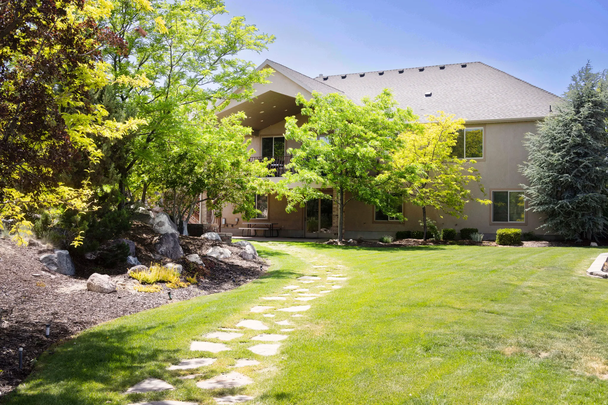 Spacious backyard featuring a stone path, mature trees, tiered landscaping with rock accents, and a covered patio with an upper-level balcony