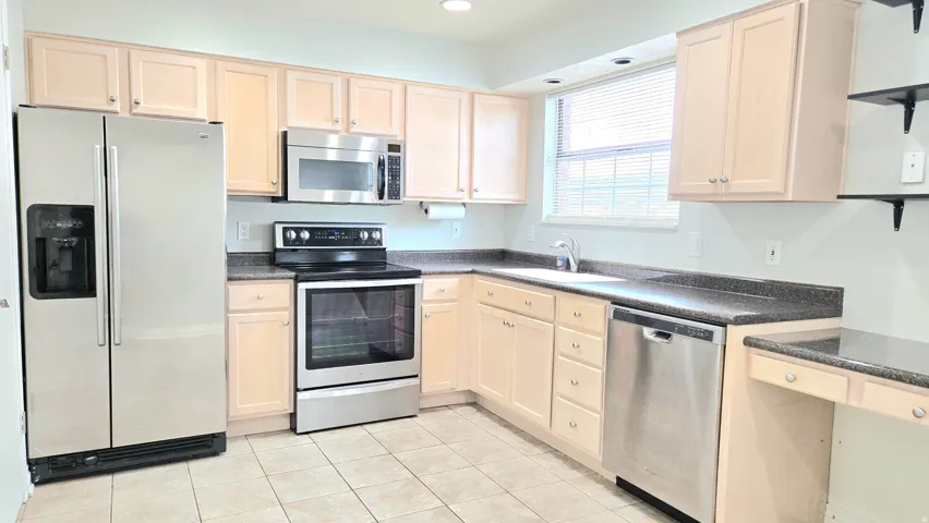 Kitchen featuring stainless steel appliances, dark countertops, light wood finish cabinetry, open shelves, and light tile patterned flooring