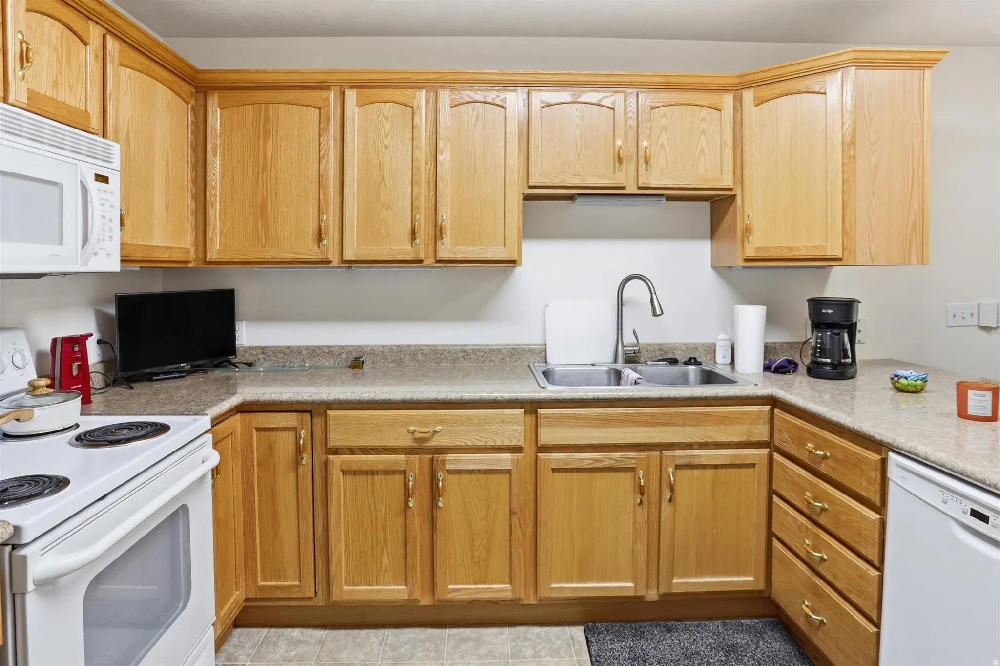Kitchen featuring light wood cabinetry with gold-tone hardware, a white built-in microwave, white electric range, and a double basin stainless steel sink with a high-arch faucet