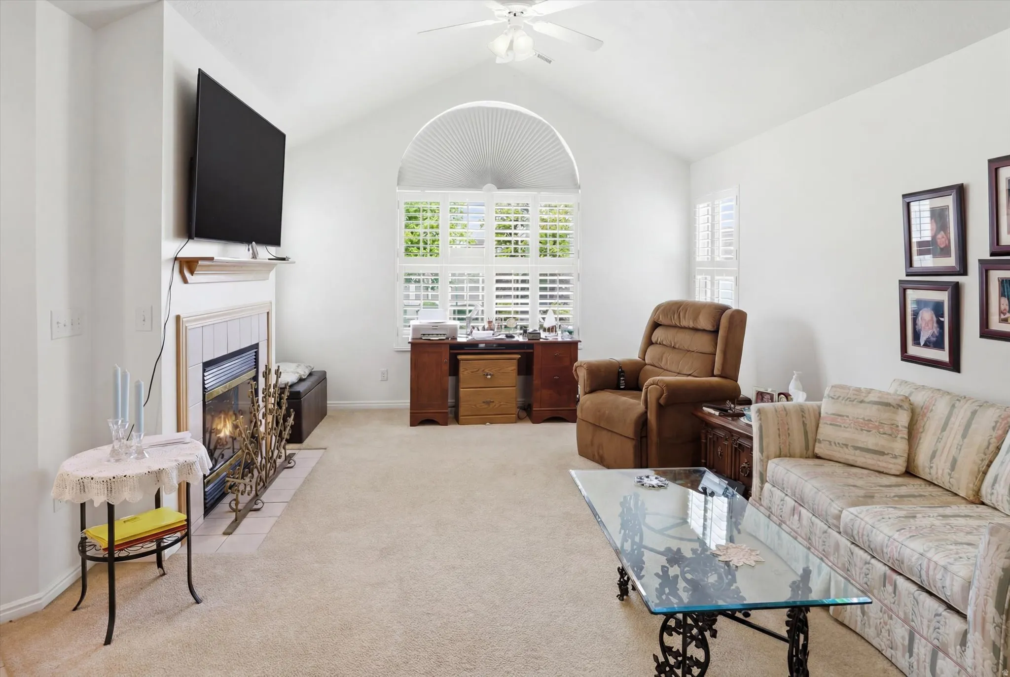 Vaulted ceiling featuring a ceiling fan, fireplace with a tile surround and mantel, and a large arched window with plantation shutters