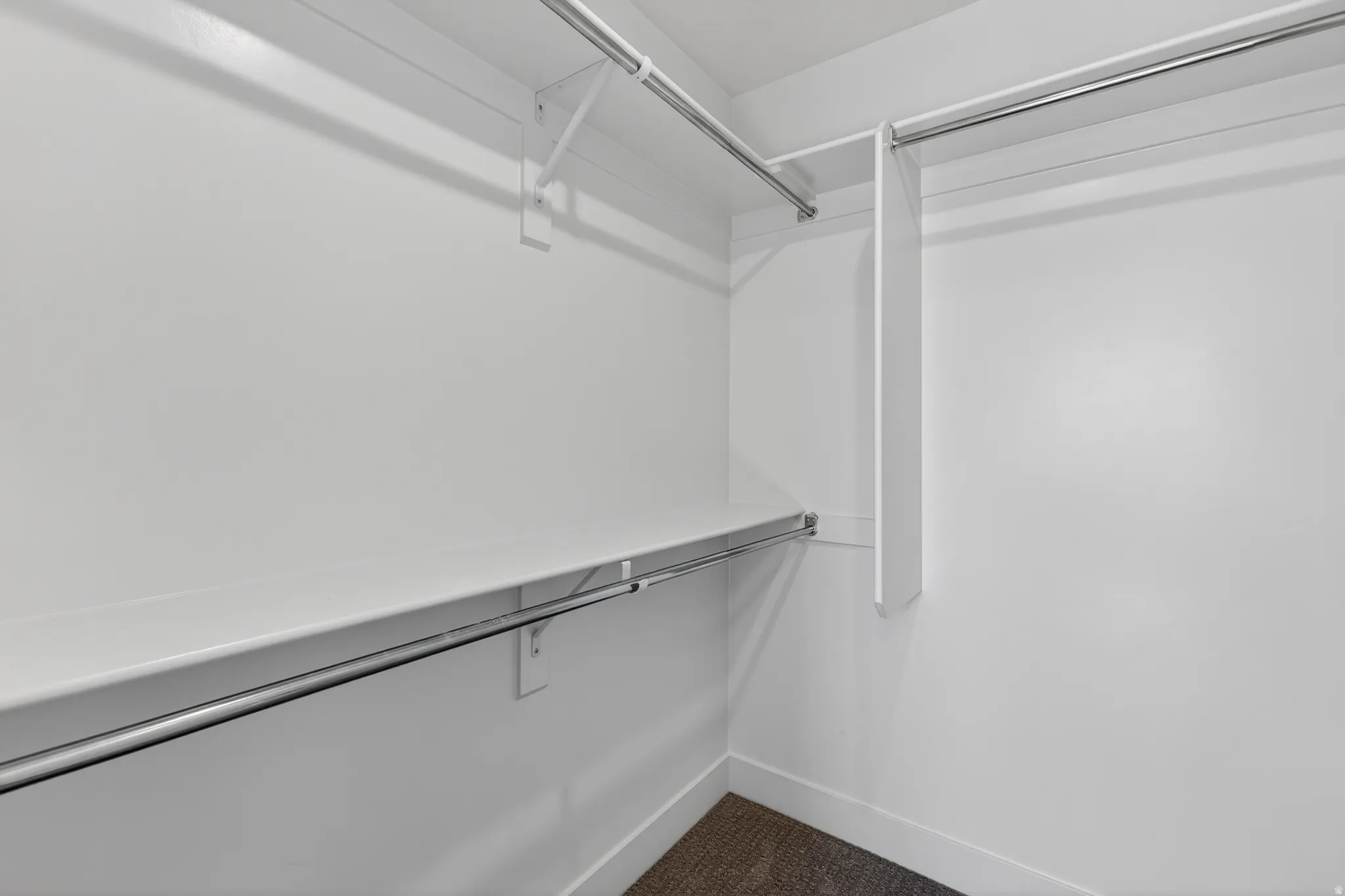 Walk-in closet with white shelving and chrome hanging rods, featuring light-colored walls and dark carpet flooring