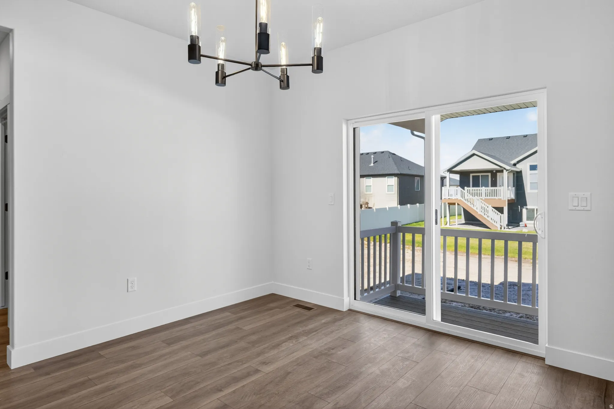 Spacious room featuring wood-finish flooring, a modern multi-bulb chandelier, white walls, and a sliding glass door providing access to a deck with white railings