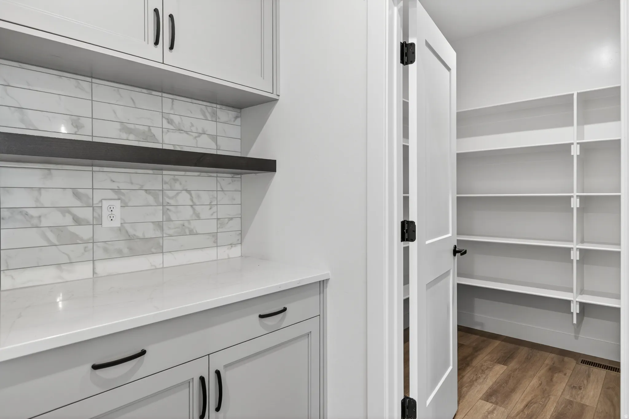 Kitchen pantry featuring light gray cabinetry, a white countertop, a floating dark wood shelf, and a white subway tile backsplash with marble-like veining