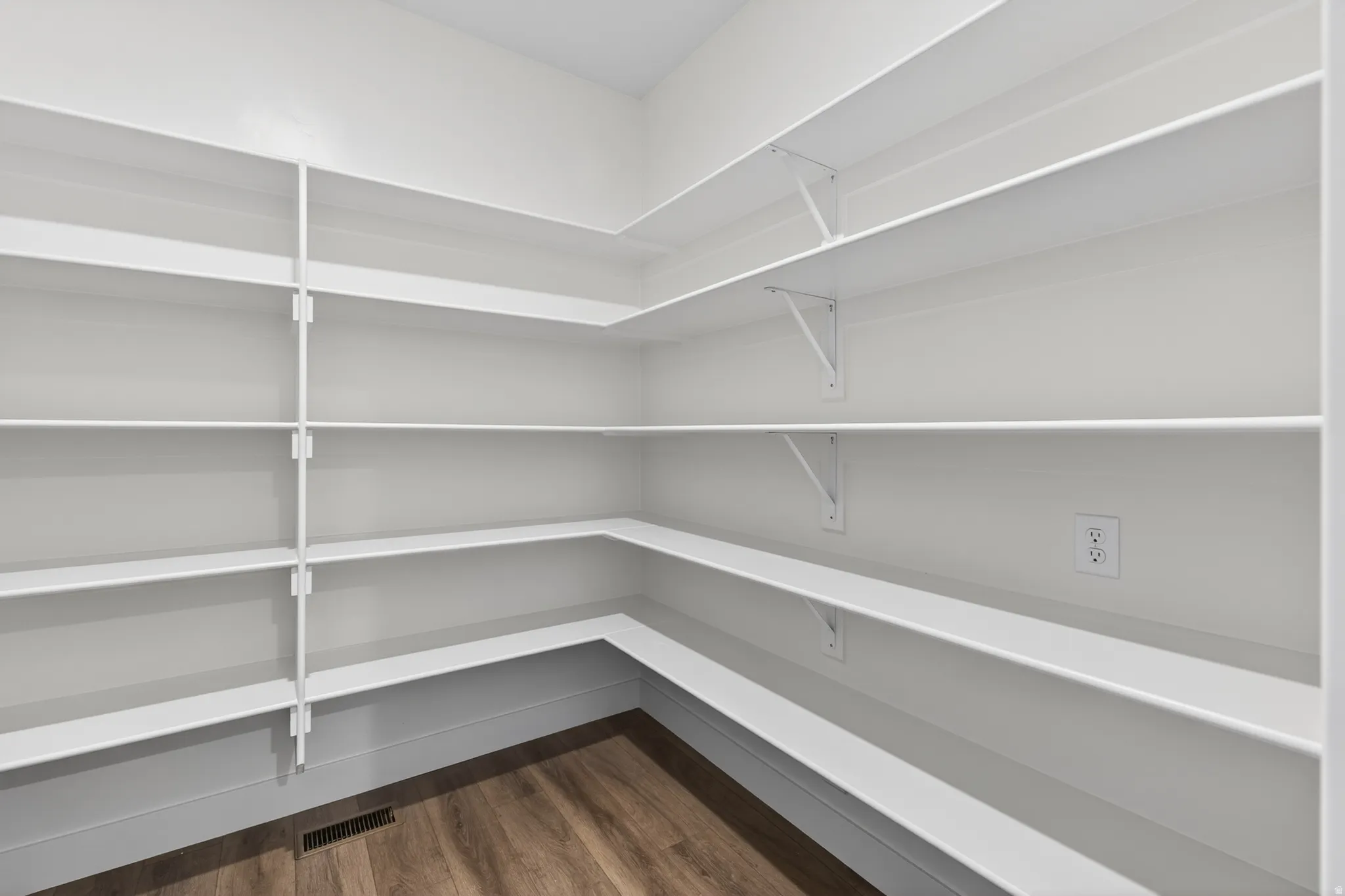 Walk-in pantry featuring extensive white shelving, wood-finish flooring, and a wall outlet