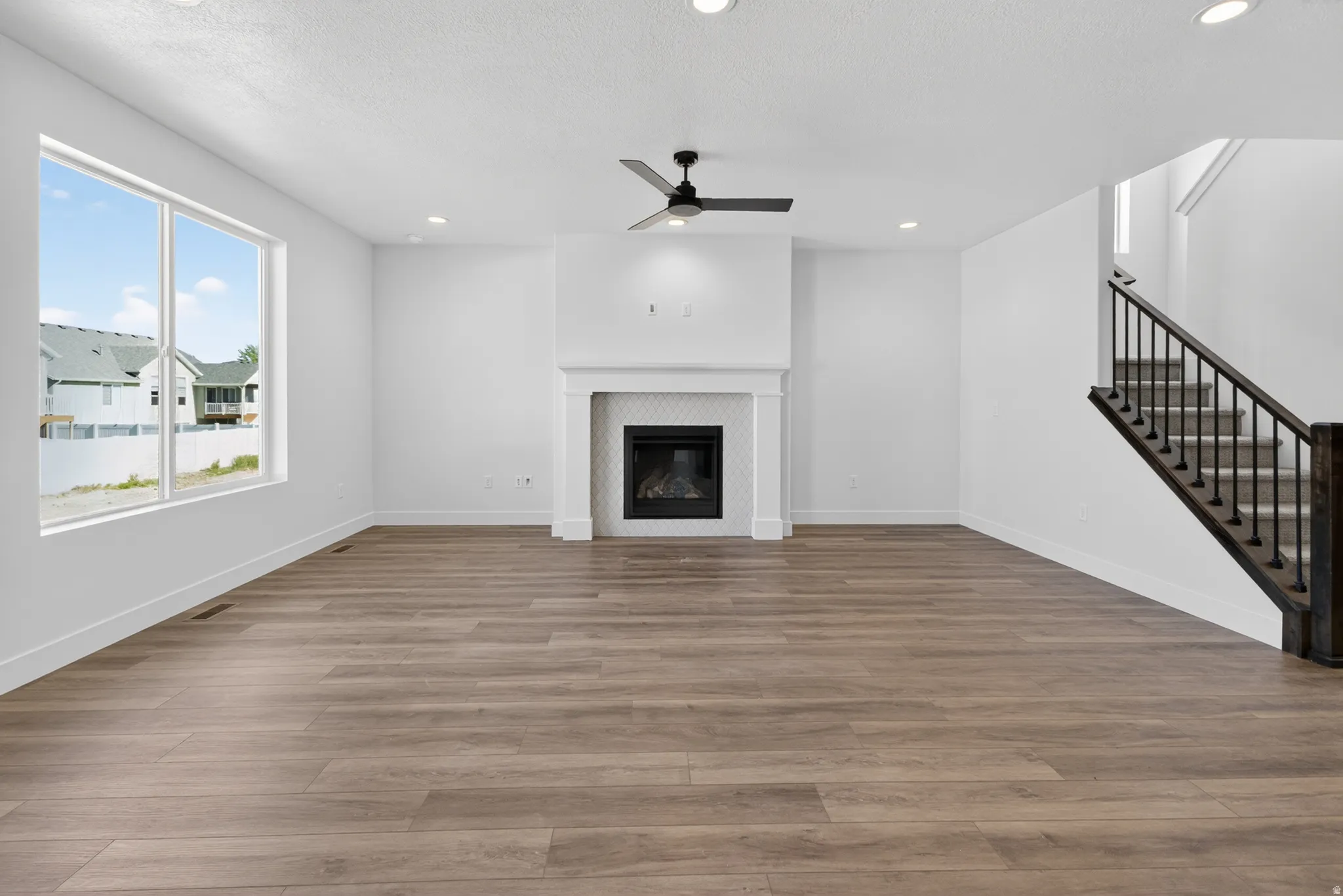 Spacious room featuring wood-finish flooring, a contemporary fireplace with a white mantel and tile surround, and a modern black ceiling fan
