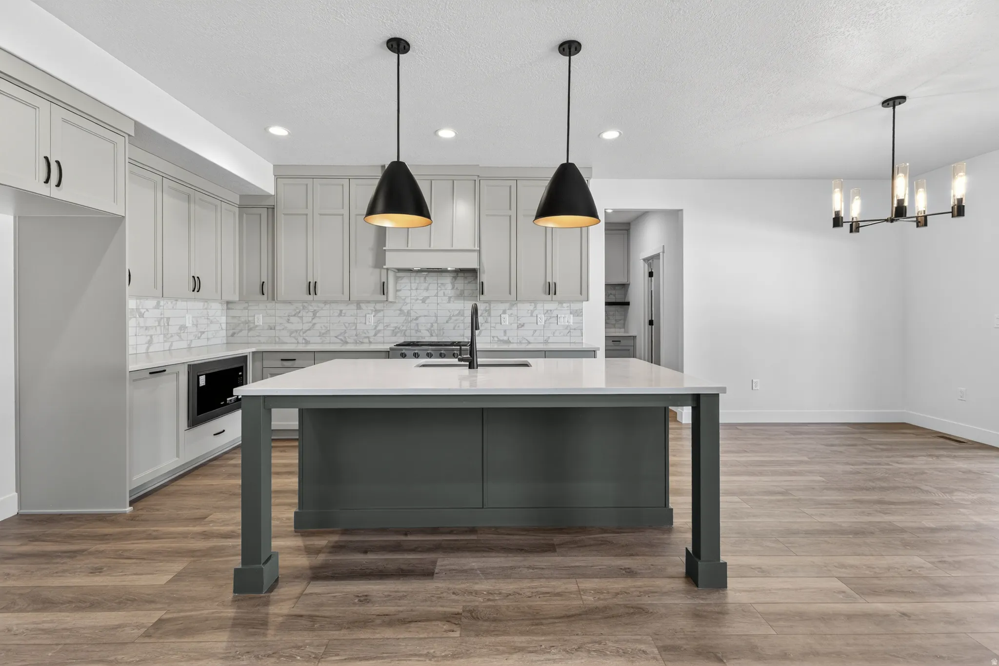 Open-concept kitchen featuring an oversized island with a light-toned countertop, gray cabinetry, a full height marble-look tile backsplash, wood-finish flooring, and black pendant lighting