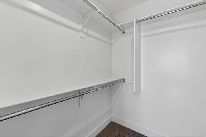 Walk-in closet with white shelving and chrome hanging rods, featuring light-colored walls and dark carpet flooring