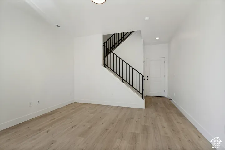 Light wood-finish flooring, white walls, black metal staircase railing, recessed lighting, and a paneled door with black hardware