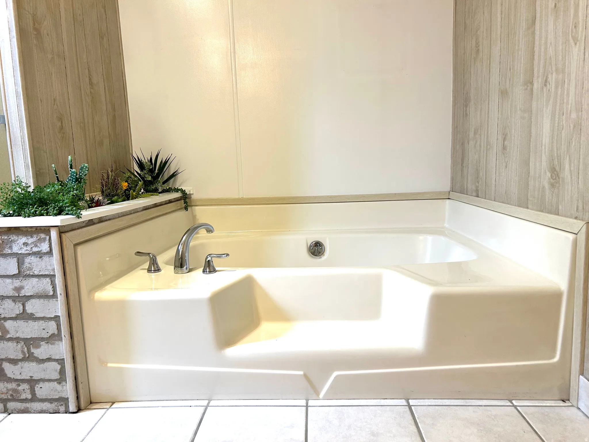 Corner soaking tub with polished chrome fixtures, surrounded by white brick-finish paneling and wood-finish wall panels, set on light tile flooring