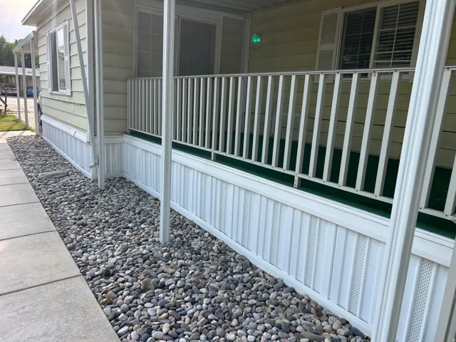 Front exterior featuring light-colored siding, a covered porch with white railings, and decorative white skirting
