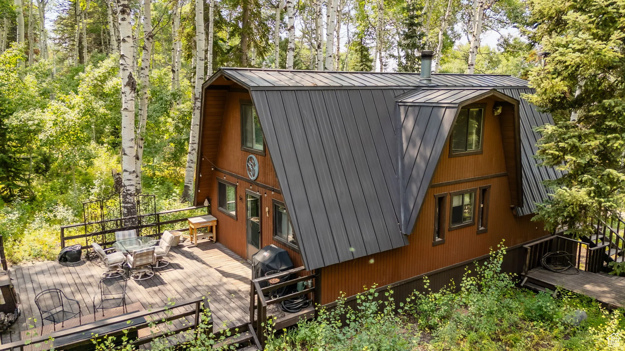 View of property exterior with a gambrel roof, outdoor dining area, a metal roof, a wooden deck, and view of scattered trees