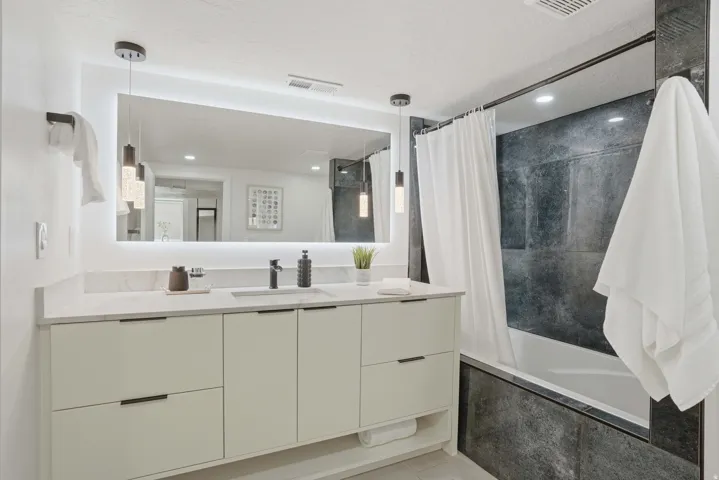 Spa like bathroom featuring vanity with back light, tiled shower / bath combo, and quartzite countertops.