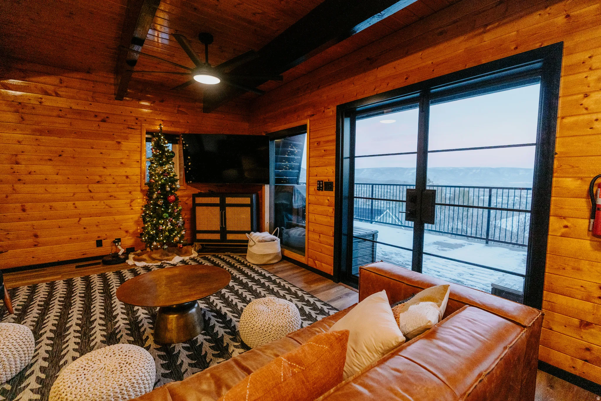 Living room with wood walls, wood finished floors, a ceiling fan, a wood ceiling with exposed beams, and a fireplace