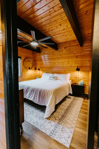Bedroom with wooden walls, a wooden ceiling with exposed beams, light wood finished floors, and ceiling fan