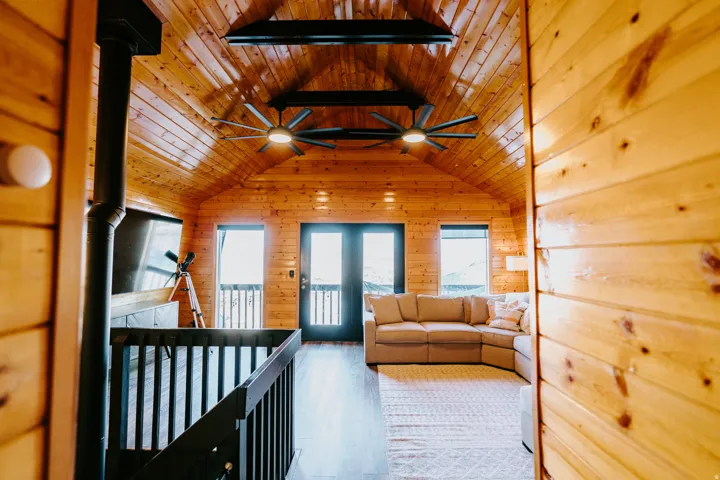 Living area with ceiling fan, light wood-style floors, wood walls, a wood ceiling with exposed beams, and a wood stove