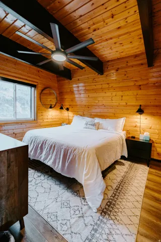 Bedroom with a wooden ceiling with exposed beams, wood walls, a ceiling fan, and wood finished floors