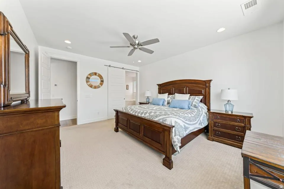 Bedroom with light carpet, a barn door, ceiling fan, recessed lighting, and ensuite bathroom