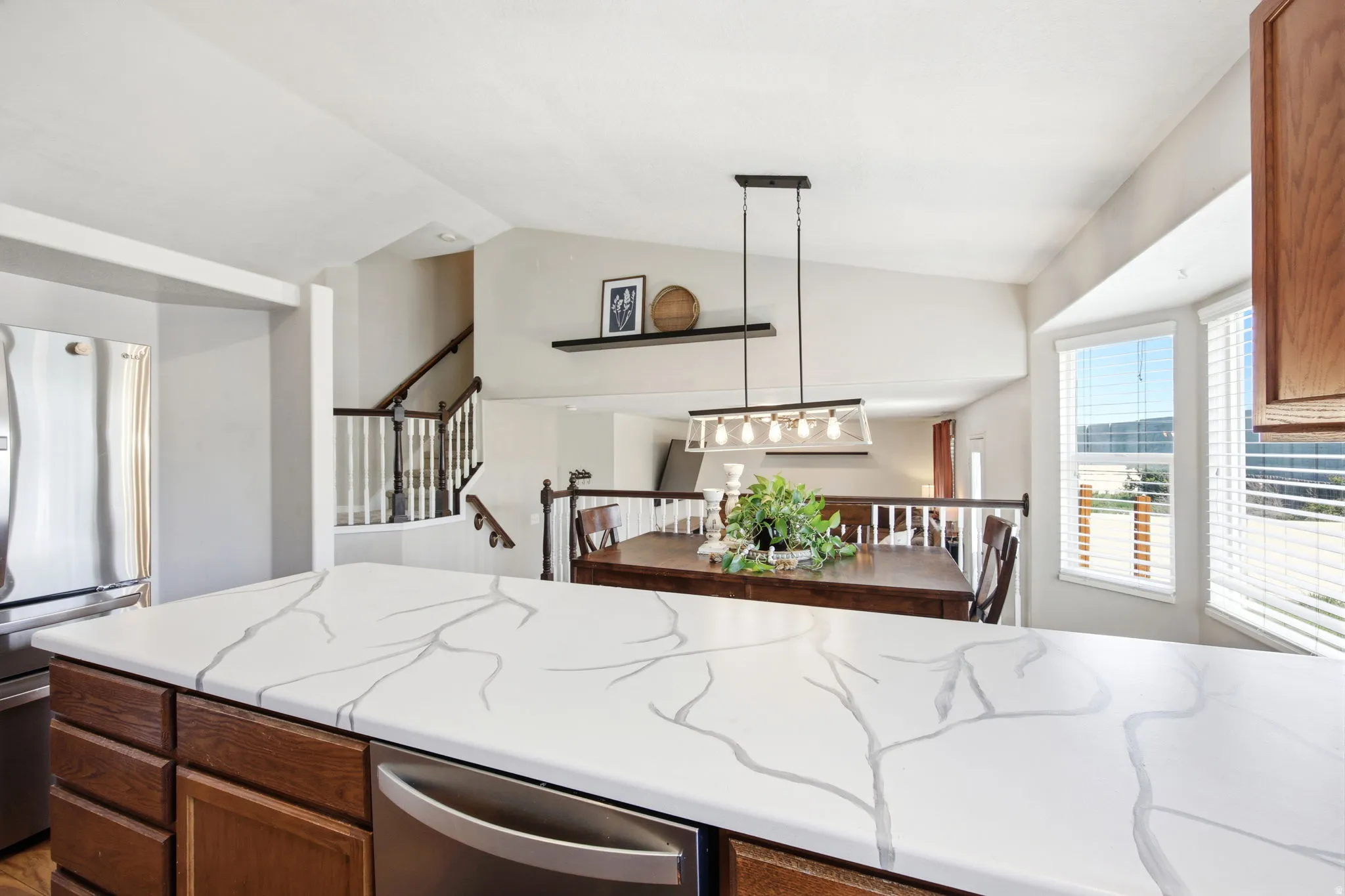 Kitchen with stainless steel appliances, lofted ceiling, decorative light fixtures, wood finish cabinetry, and light stone countertops