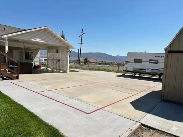 Expansive concrete patio area with painted court lines, featuring a covered carport and a detached storage shed