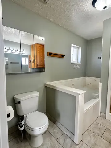 Bathroom featuring a built-in jetted tub, ceramic tile flooring, a white toilet with bidet attachment, a mirrored medicine cabinet, and a small window