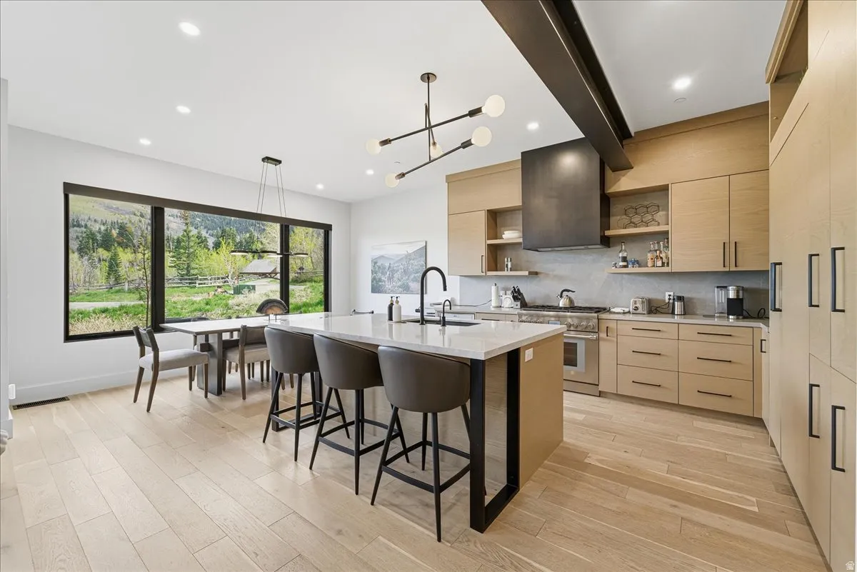 Kitchen with open shelves, light wood finish cabinetry, a center island with sink, light stone countertops, and beam ceiling