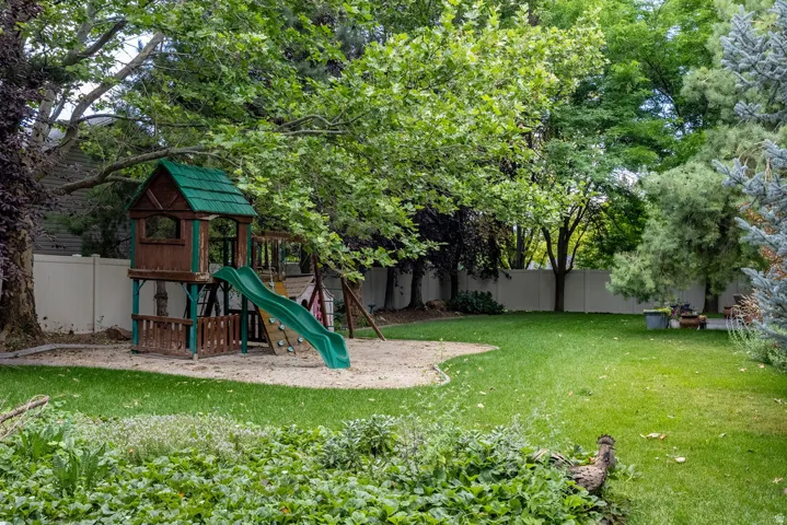 Expansive backyard featuring a lush lawn, mature trees providing ample shade, a sand play area with a built-in playset, and a private perimeter fence