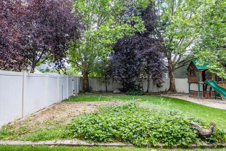 Expansive backyard featuring a white vinyl privacy fence, mature trees, a wooden play structure with a green slide, and varied ground cover