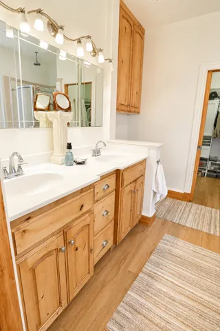 Bathroom vanity featuring dual undermount sinks, white countertops, natural wood cabinetry, brushed nickel hardware, and wood-finish flooring
