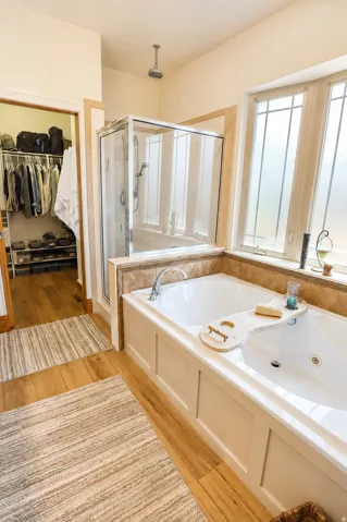 Soaking tub with a paneled surround, glass-enclosed shower, wood-finish flooring, and a large window with frosted panes