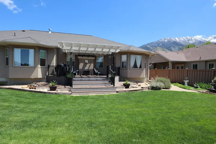 Rear exterior featuring a multi-level deck with pergola, bay windows, a stone-lined garden bed, and a spacious lawn with mountain backdrop