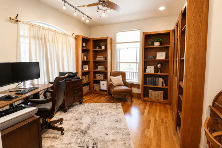 Hardwood flooring throughout featuring extensive built-in shelving, an arched window, a track lighting system, and a ceiling fan