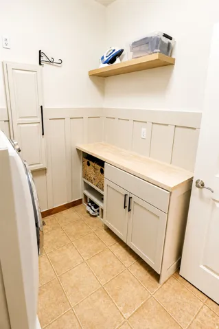 Laundry area featuring beige square tile flooring, a custom built-in cabinet with a wood-finish countertop, a floating wood-finish shelf, and board and batten wall detailing