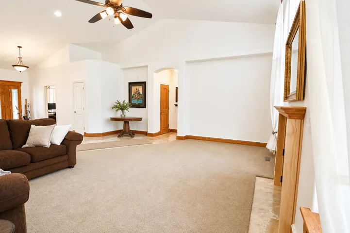 Spacious living area featuring vaulted ceilings with a ceiling fan, carpet flooring, a stone hearth, and natural wood trim throughout
