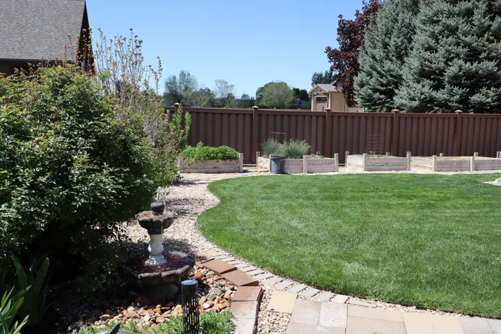 Expansive lawn area featuring a stone water fountain, gravel pathway, mature evergreen trees, and raised garden beds