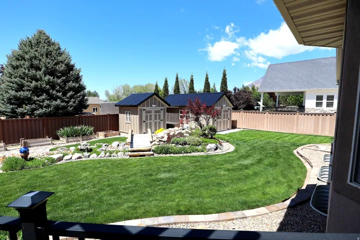 Expansive backyard featuring a lush lawn, two storage sheds with dark roofs, a decorative rock garden with plantings, and a full privacy fence