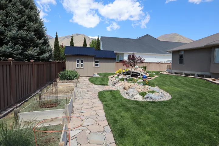 Manicured backyard with a flagstone pathway, a decorative rock garden with a small bridge feature, a detached storage shed with a dark metal roof, and raised garden beds