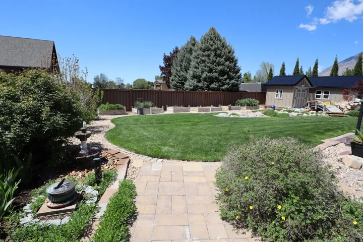 Expansive backyard featuring a lush lawn, paver stone walkway, privacy fencing, custom planter boxes, and a storage shed with a dark metal roof