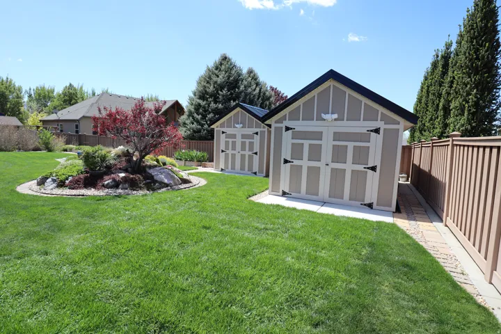 Expansive green lawn featuring two gable-roof storage sheds, a decorative rock garden with a red-leaf tree, a tan privacy fence, and a paver stone walkway