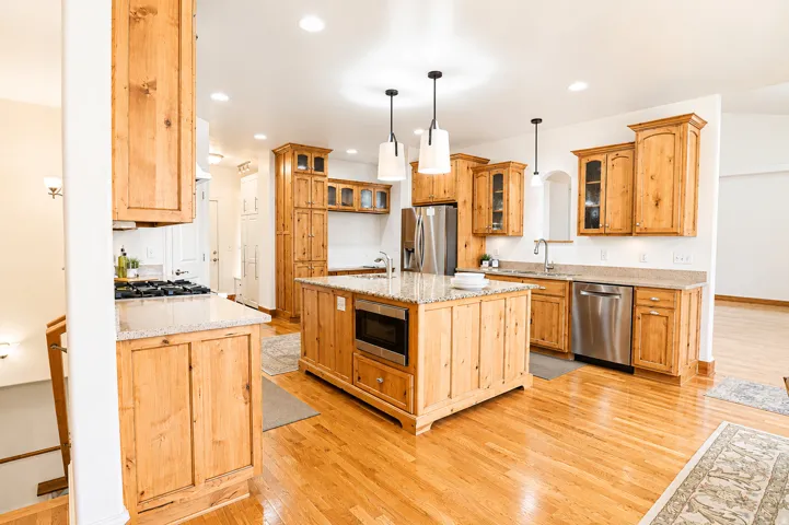 Spacious kitchen featuring light wood cabinetry, stone countertops, stainless steel appliances, a central island with a built-in microwave, and wood flooring