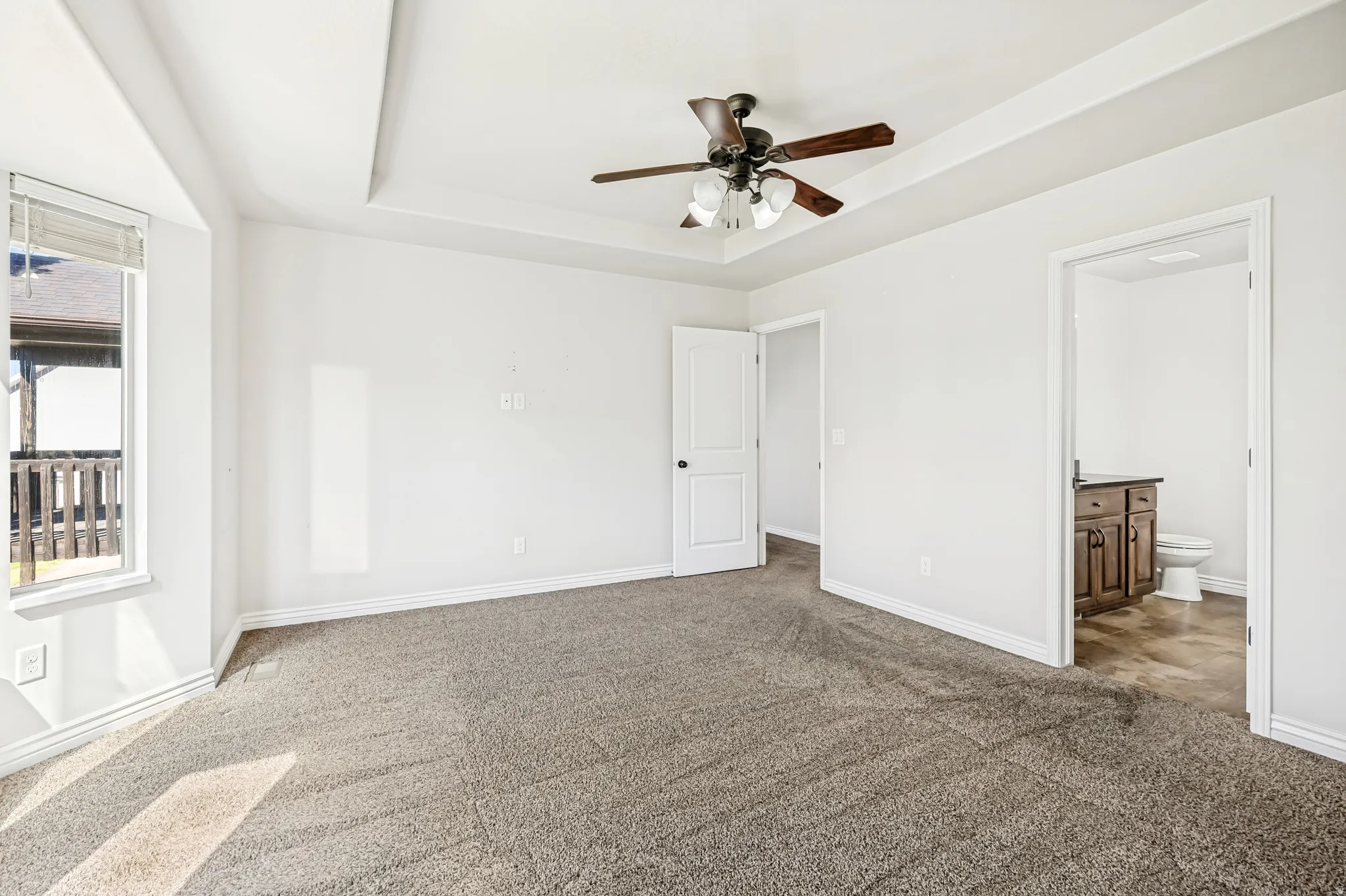 Unfurnished bedroom with dark colored carpet, a raised ceiling, connected bathroom, and ceiling fan