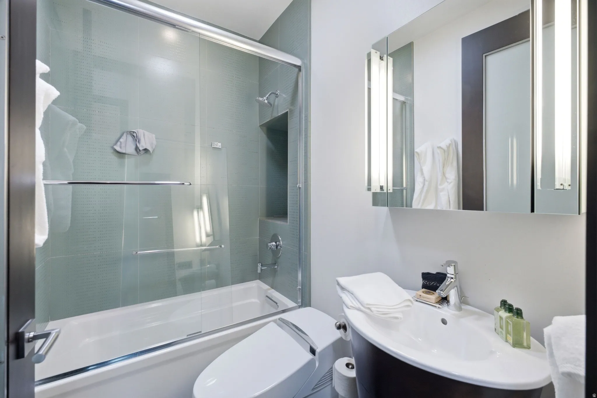 Modern bathroom featuring a built-in tub with glass sliding doors, integrated shower niche, a wall-mounted medicine cabinet with integrated lighting, and a contemporary vanity with an undermount sink